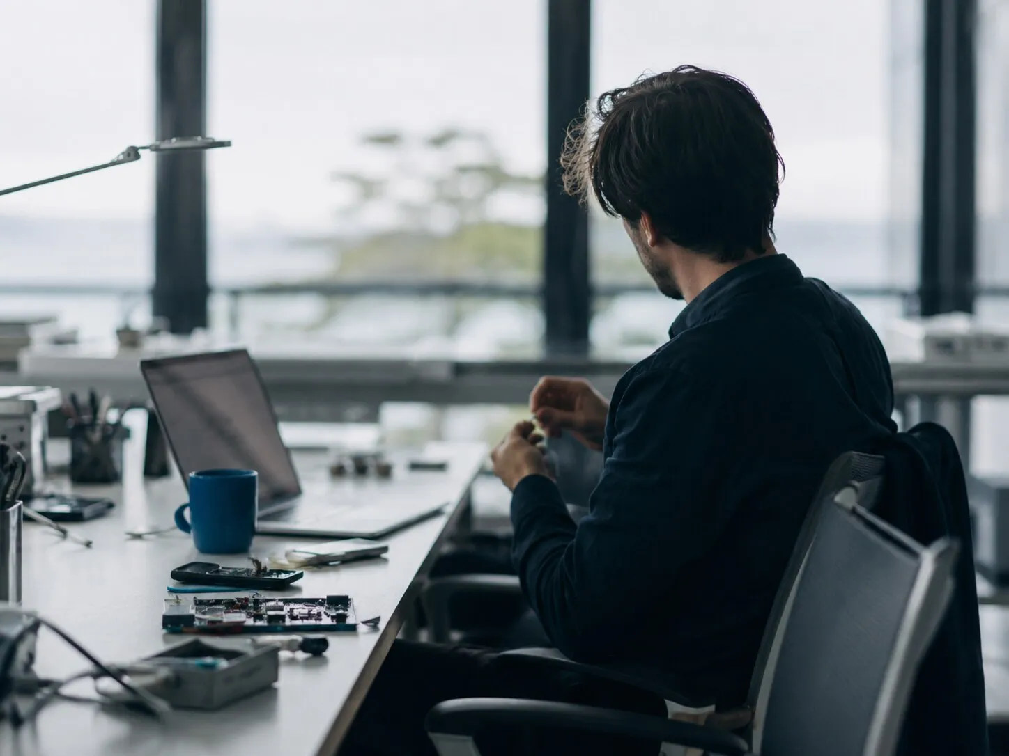 An engineer at a desk of half-assembled hardware, working by a window over the water.